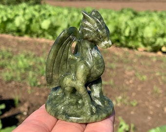 Op de afbeelding: Gedetailleerd groen drakenbeeldje met vleugels en een gedetailleerd lichaam. De draak is gesneden uit een steenachtig materiaal en staat op een ronde basis. Het beeldje is een decoratief item.
