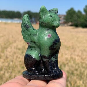 May include: A carved stone cat figurine with green and black coloring, featuring red accents. The cat has a seated pose with its tail curled and wings. The figurine is held in a hand against a blurred outdoor background.