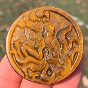 May include: A round, amber-colored pendant with a carved relief of a reclining figure with long flowing hair. The figure is surrounded by stylized waves and clouds. The pendant is held in a person's hand.