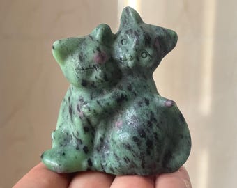 May include: A carved stone sculpture of two cats embracing. The cats are green with black speckles and small pink accents. The sculpture is held in a hand and has a smooth, polished finish.