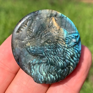 May include: A round, carved labradorite stone featuring a detailed wolf head. The stone has a mix of blue, green, and gray hues, with the wolf's fur and features intricately carved. The background is blurred, suggesting an outdoor setting.