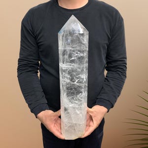 May include: A large, clear quartz crystal point with a hexagonal shape and a pointed top. The crystal features internal fractures and natural inclusions throughout its transparent body, showcasing a polished surface and a solid, heavy appearance.