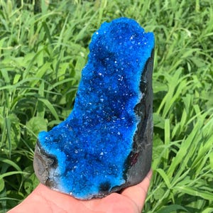 May include: A vibrant blue crystal geode specimen featuring a cluster of small, sparkling, deep blue crystalline structures on the interior surface, contrasted by a dark, rough, and textured exterior rock formation.