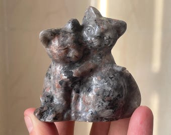 May include: A stone sculpture of two cats embracing. The cats are gray with black and pink speckles. One cat is hugging the other from behind. The sculpture is held in a person's hand.
