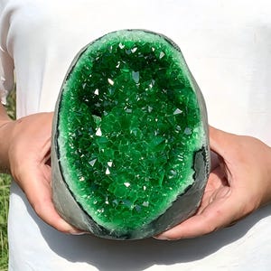 Op de afbeelding: Een grote, ovaalvormige geode met een diep, levendig smaragdgroen kristallijn interieur. De buitenkant van de steen is donkergrijs en mat, wat contrasteert met de gefacetteerde, doorschijnende groene kristallen die de holle kern vullen.