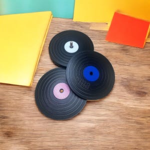 以下が含まれることがあります： 白、青、ピンクのカラーセンターラベルが付いた、3つの黒いビニールレコードコースター。コースターは木製の表面に配置され、背景にはカラフルな紙があります。