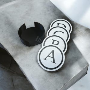 Puede incluir: Un juego de cuatro posavasos blancos y negros con las letras A, B, C y D impresas. Los posavasos son redondos con un borde negro. Un portavasos negro está al lado.