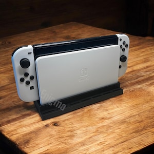 以下が含まれることがあります： 白のNintendo Switch本体と白いコントローラーが黒いスタンドにドッキングされています。スタンドには「Enigma」の文字が印刷されています。コンソールは木製の表面にあります。