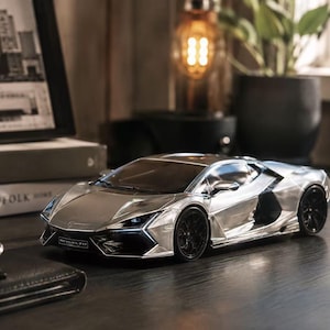 May include: A silver Lamborghini Revuelto scale model car featuring a sharp, angular body design, black multi-spoke wheels, and a prominent front bumper with the Revuelto nameplate clearly visible on the front grille area of the vehicle.