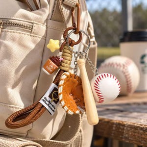 May include: A keychain featuring a brown paracord strap with a white tag, attached to a metal ring with charms including a yellow star, a brown coffee cup, a tan baseball glove, a wooden baseball bat, and a white baseball with red stitching.