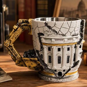 Könnte beinhalten: Ein Keramikbecher mit einem weißen Gebäude mit Säulen und einer rissigen Außenstruktur. Der Henkel ist wie ein goldfarbener mechanischer Baggerarm geformt, an dessen Seite sich eine mit schwarzem Material gefüllte Baggerschaufel befindet.