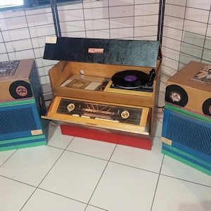 Peut inclure: Une console tourne-disque en bois de style vintage avec un couvercle noir, comprenant un tuner radio central avec des boutons dorés, flanqué de deux haut-parleurs assortis. Les enceintes ont des bases bleu et vert, des dessus beige et des grilles.