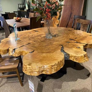May include: A large, unique wooden table with a natural edge, showcasing a light brown color and a distinctive, organic shape. The table is supported by a black metal base and is surrounded by wooden chairs. A vase of flowers sits on top.