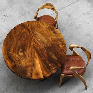 May include: A circular wooden table with a dark brown, patterned surface. Two chairs with curved wooden frames and brown leather seats are arranged around the table. The chairs have a distinctive, artistic design.
