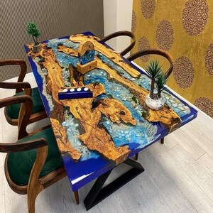 Pode incluir: Uma mesa de jantar retangular com um design de rio em resina epóxi azul, com elementos de madeira natural e detalhes decorativos. A mesa é suportada por pernas de metal preto e rodeada por cadeiras de madeira com assentos de veludo verde.