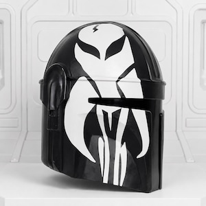 May include: Black helmet with a white graphic design. The helmet features a stylized white emblem on the front and top, with a lightning bolt symbol. The helmet is likely a costume piece or collectible.
