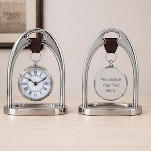 May include: Two silver-toned desk clocks with a classic design. One clock displays the time, while the other has space for personalization. Each clock is suspended within a decorative arch and has a brown leather strap.