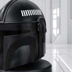 May include: A black helmet with a matte finish, featuring a series of white vertical lines on the upper right side. The helmet has a detailed design with a visor and a rounded shape, set against a bright, white background. The helmet is likely a collectible or cosplay item.