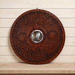 Op de afbeelding: Een rond houten schild met ingewikkelde handgesneden knoopmotieven, een centrale metalen naaf en runeninscripties langs de buitenrand. Het donkerbruine hout toont gedetailleerde reliëfsnijwerken van een boom en cirkelvormige symbolen.
