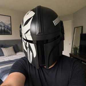 May include: A black and white Mandalorian helmet with a glossy finish. The helmet features a distinctive design with white stripes and accents, and a visor. The helmet is a replica of the one worn by the character in the Star Wars series.