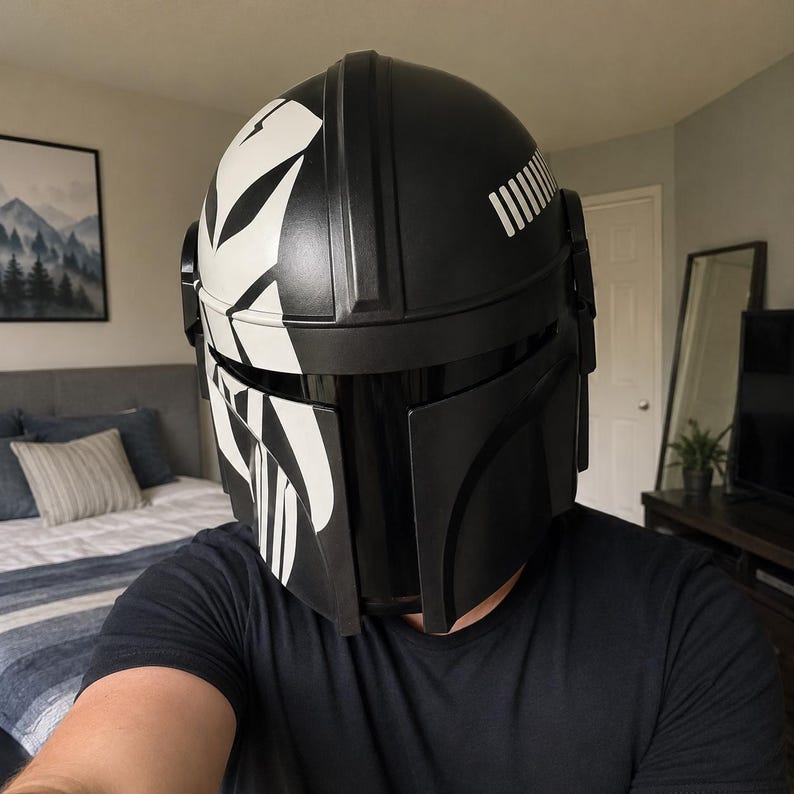 May include: A black and white Mandalorian helmet with a glossy finish. The helmet features a distinctive design with white stripes and accents, and a visor. The helmet is a replica of the one worn by the character in the Star Wars series.