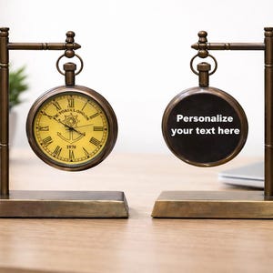 May include: Two antique-style pocket watch clocks on stands. One clock has a yellow face with Roman numerals and the text "Victoria London". The other clock has a black face with the text "Personalize your text here".