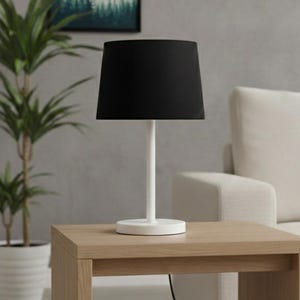 May include: A modern table lamp with a black lampshade and a white base. The lamp sits on a light-colored wooden side table. A potted plant is in the background, and a white sofa is to the right.