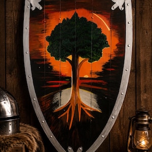 Op de afbeelding: Een houten schild met een geschilderde afbeelding van een grote groene boom met zichtbare wortels tegen een levendige oranje en zwarte zonsondergang. Het schild heeft een zilverkleurige rand die is vastgezet met gelijkmatig verdeelde klinknagels.