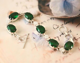 May include: A silver bracelet featuring oval-cut, emerald-green gemstones. The bracelet has a toggle clasp and is accented with small, clear stones between the larger gems. The jewelry is displayed on a soft, patterned background.