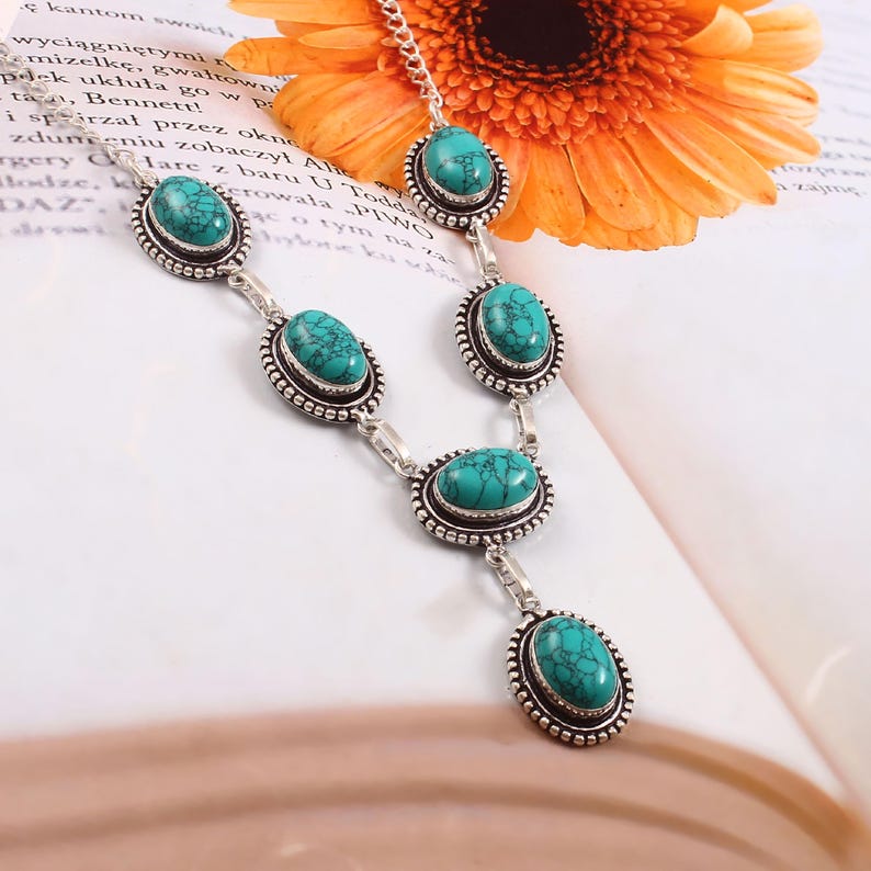 May include: A silver necklace with oval turquoise gemstones. Each stone is set in a beaded frame. The necklace is displayed on a white surface with a partially visible orange flower and text.