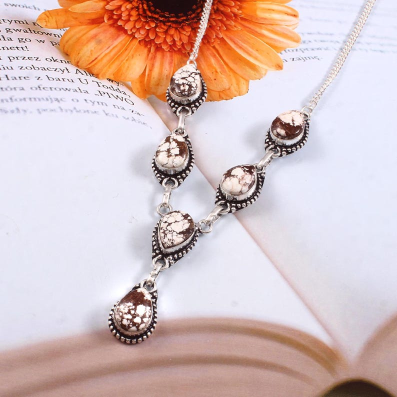 Puede incluir: Un collar plateado con colgantes en forma de l&aacute;grima con un patr&oacute;n moteado blanco y marr&oacute;n. El collar tiene una cadena delicada y se muestra sobre un libro con una flor en el fondo.