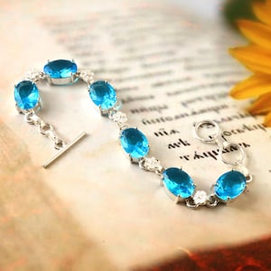 Puede incluir: Una pulsera de plata con piedras preciosas ovaladas de color azul brillante. La pulsera tiene un cierre de palanca y está engastada con piedras más pequeñas y transparentes entre las gemas azules más grandes. La joya se exhibe sobre un fondo de estilo vintage.