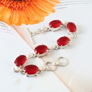 Può includere: Bracciale in argento con pietre preziose ovali rosso intenso. Il bracciale ha una chiusura a levetta. Le pietre preziose sono incastonate in metallo argentato. Sfondo morbido e neutro, fiore arancione brillante.
