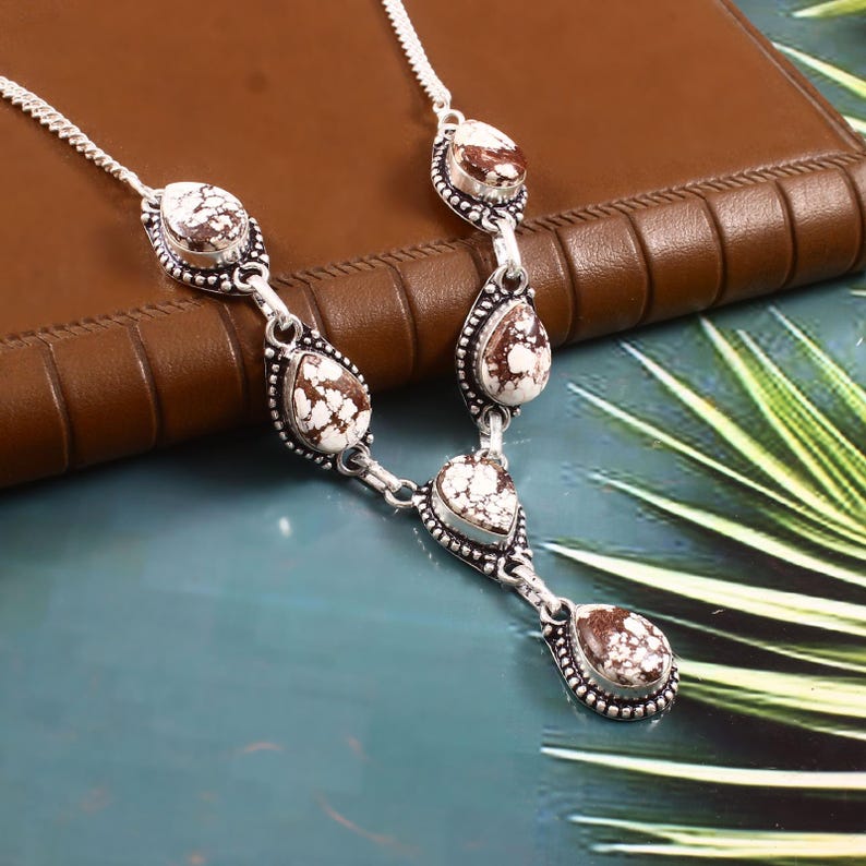 Puede incluir: Un collar plateado con piedras blancas y marrones estampadas en forma de l&aacute;grima y ovalada. Las piedras est&aacute;n engarzadas en un marco detallado y con cuentas, conectadas por eslabones plateados. El collar descansa sobre una superficie de cuero marr&oacute;n.