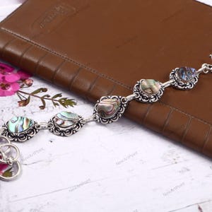 Può includere: Un bracciale in argento con cinque maglie a forma di cuore in conchiglia di abalone. I gusci iridescenti sono incastonati in cornici d'argento ornate. Il bracciale è su una superficie di pelle marrone.