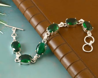 May include: A silver bracelet featuring oval-cut, emerald-green gemstones. The bracelet has a toggle clasp and is displayed on a brown leather surface. The gemstones are set in a silver chain.