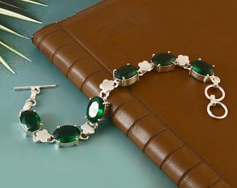 May include: A silver bracelet featuring oval-cut, emerald-green gemstones. The bracelet has a delicate design with a toggle clasp. The gemstones are set in a silver metal setting. The bracelet is displayed on a brown leather surface.