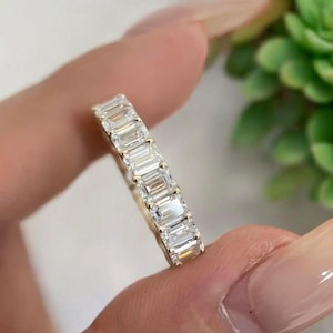 May include: A gold eternity band featuring a continuous row of clear, rectangular emerald-cut stones set in a shared prong setting. The ring displays a polished metal finish and a series of faceted, transparent gemstones arranged in a straight line.