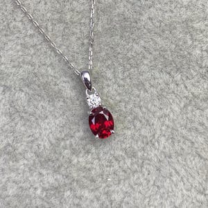 May include: A silver necklace with a pendant featuring a large, oval-cut, deep red gemstone. Above the red stone is a smaller, round, clear gemstone. The necklace chain is delicate and silver.