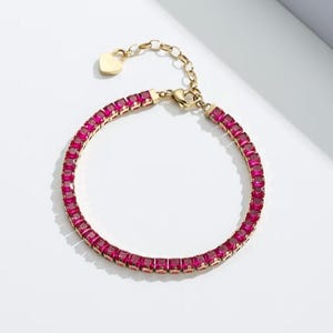 May include: Gold-toned bracelet featuring a row of square, ruby-colored gemstones. The bracelet has a delicate chain and a heart-shaped charm near the clasp. The gemstones are set closely together, creating a continuous line of color.