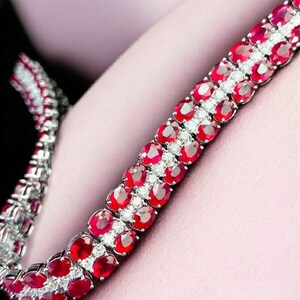 May include: A close-up of a silver bracelet with two rows of oval-cut, vibrant red gemstones. The gemstones are interspersed with small, clear stones, creating a sparkling effect. The bracelet is set against a soft pink background.