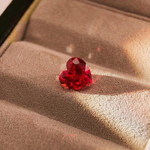 以下が含まれることがあります： ハート型で深紅色のファセット宝石。 宝石は、暗い箱の中のニュートラルな色調のテクスチャ加工された表面に置かれ、印象的なコントラストを生み出しています。 照明は宝石の輝きと形を際立たせています。