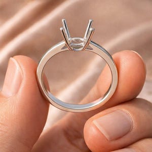May include: A silver ring setting with prongs designed to hold a gemstone. The ring is held between fingers, showcasing its polished band and the open setting ready for a stone. The ring is a jewelry item.