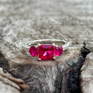 May include: A silver ring featuring a large, rectangular, bright pink gemstone flanked by two smaller, heart-shaped pink gemstones. The ring band is detailed with small, clear stones. The ring is set against a textured, wooden surface.