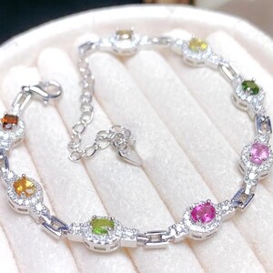 May include: A silver bracelet featuring a series of colorful gemstones, including orange, yellow, green, and pink, each surrounded by small, clear accent stones. The bracelet has a delicate chain design and a secure clasp.