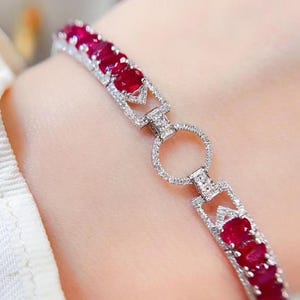 May include: A silver-toned bracelet featuring a series of oval-cut deep red rubies set in metal frames. The design includes a central circular link encrusted with small clear stones, connecting two segments of rubies and pavé-set metal links.