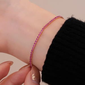 May include: A gold-toned bracelet with a continuous line of small, bright pink gemstones. The bracelet is worn on a wrist, with a black sleeve in the background. The gemstones are closely set, creating a sparkling effect.