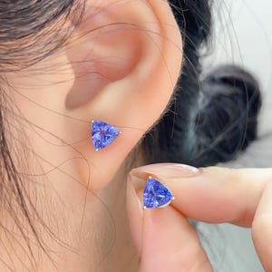 May include: A pair of blue triangular gemstone stud earrings. One earring is in the ear, and the other is held up by a hand. The earrings have a silver setting and a vibrant blue color.