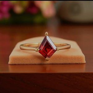 May include: A gold ring with a large, square-cut, deep red gemstone. The gemstone is set in a diamond shape, and the ring band is a simple, thin gold band. The ring is displayed on a small, tan velvet surface.