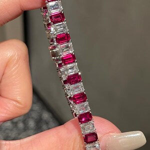 May include: A tennis bracelet featuring alternating emerald-cut deep red rubies and clear white diamonds set in a silver-toned metal band. The stones are arranged in a repeating pattern along the length of the bracelet, showcasing a polished finish.